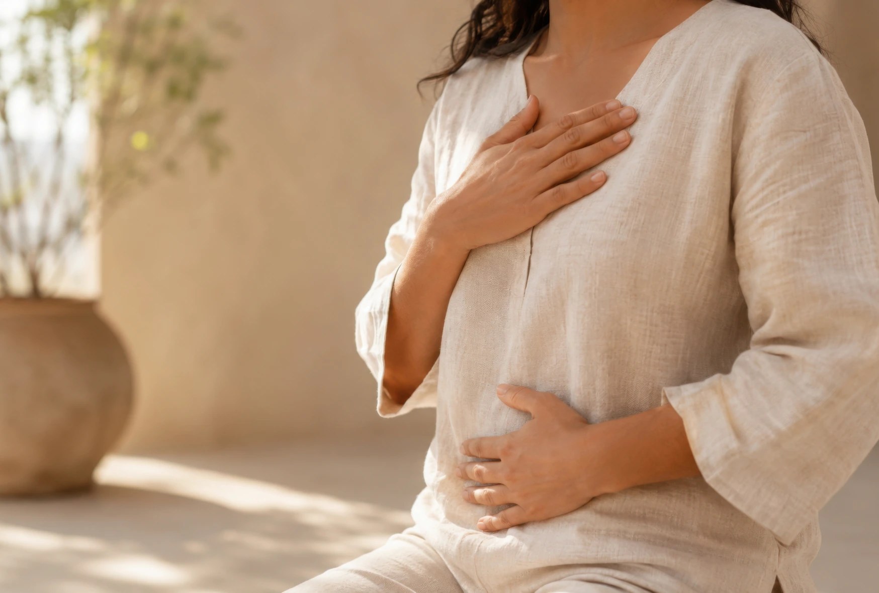 Woman in a soft cream top resting one hand on her heart and one on her belly in warm natural light